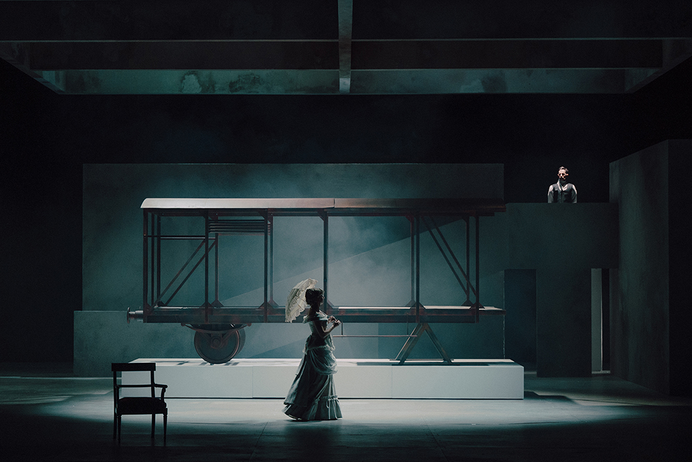 fotos ze spektaklu przedstawia damę z parasolką na tle industrialnego wozu