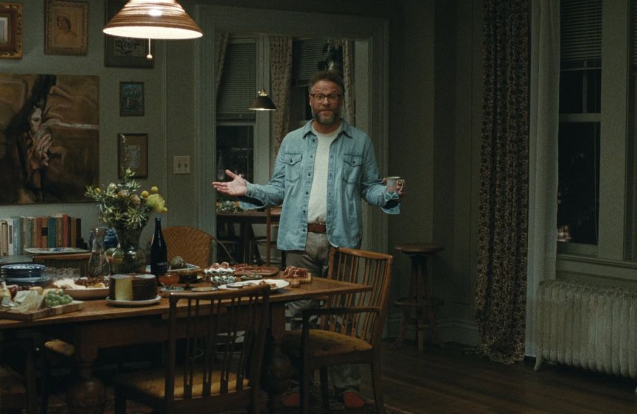 Kadr z filmu „Zaproszenie”. Postawny mężczyzna (Seth Rogen) z brodą, w jeansowej koszuli i okularach stoi obok stołu z przekąskami. Rozkłada ręce w zapraszającym geście, ale ma zirytowaną minę.